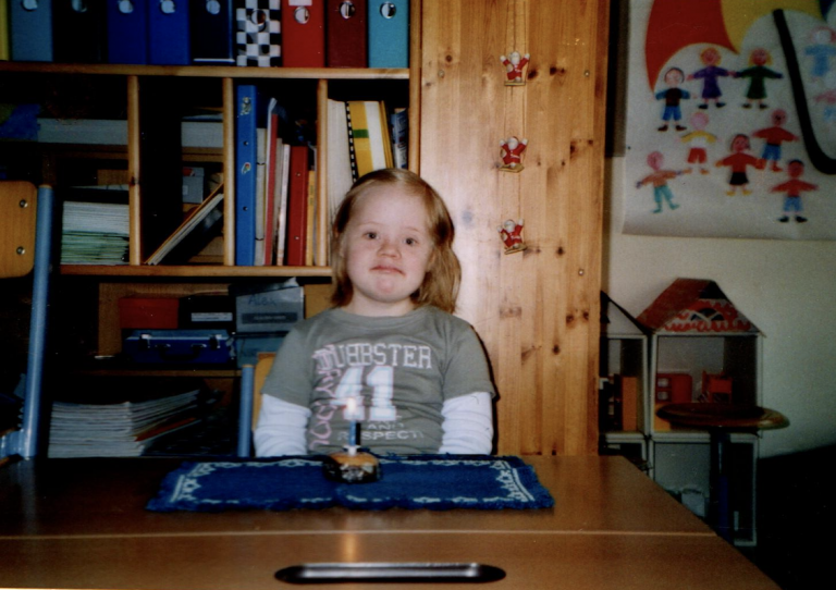 Die Autorin Hanna Gugler sitzt als Volksschülerin vor einem Holzregal. Im regal sind Mappen und Bücher.
