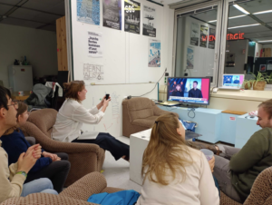 Foto aus unserem Büro: Lisa, Luise, Artin, Clara und Lukas sitzen vor einem Fernsehbildschirm. Zu sehen der Live-Stream von Puls24 mit Kathi und Sebastian im Bild. Lisa fotografiert den Bildschirm. Alle anderen schauen auf den Fernseher.