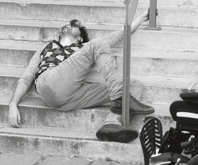Ein Mann liegt auf den Stufen einer Treppe. Vorne im Bild ist ein Teil seines Rollstuhls zu sehen.