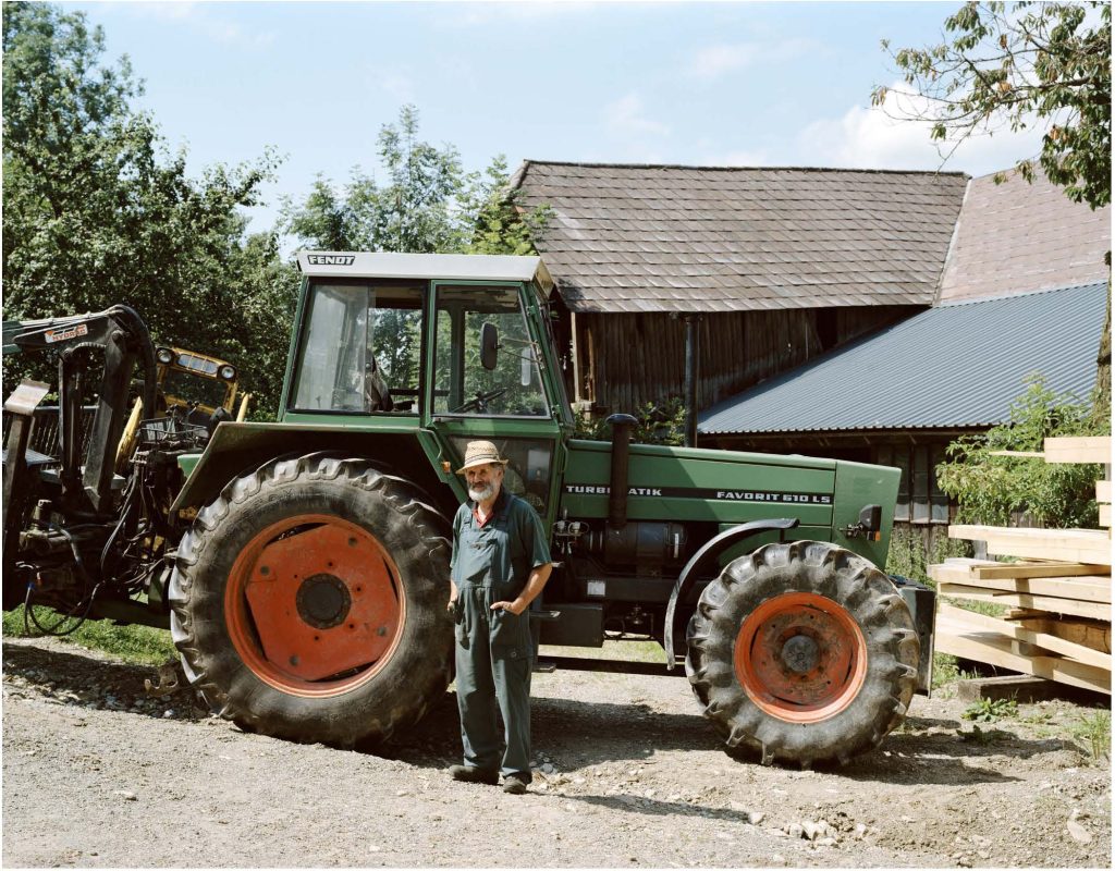 Ein älterer Mann steht vor einem grünen Traktor. Er trägt einen Hut und schaut mit ernsten Blick in die Kamera.