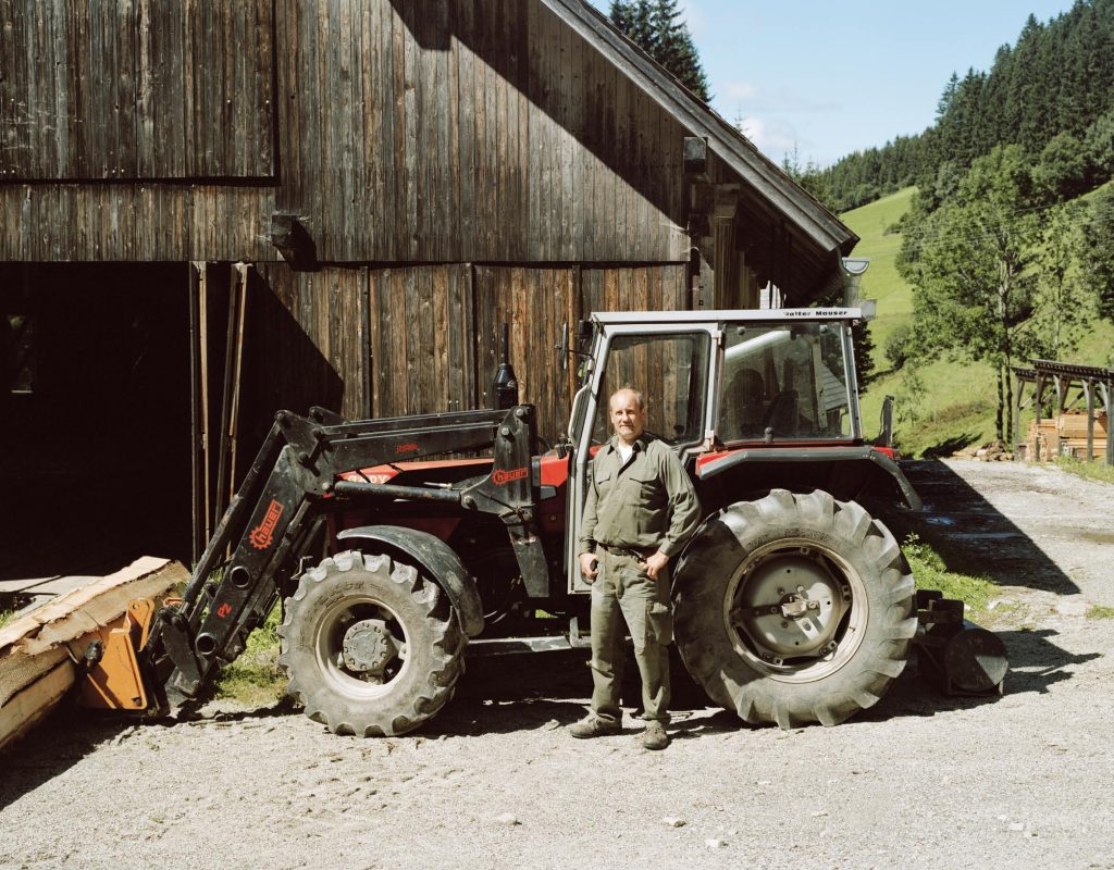 Ein älterer Mann steht vor einem Traktor. Er schaut mit ernsten Blick in die Kamera. Im Hintergrund sieht man eine Scheune.
