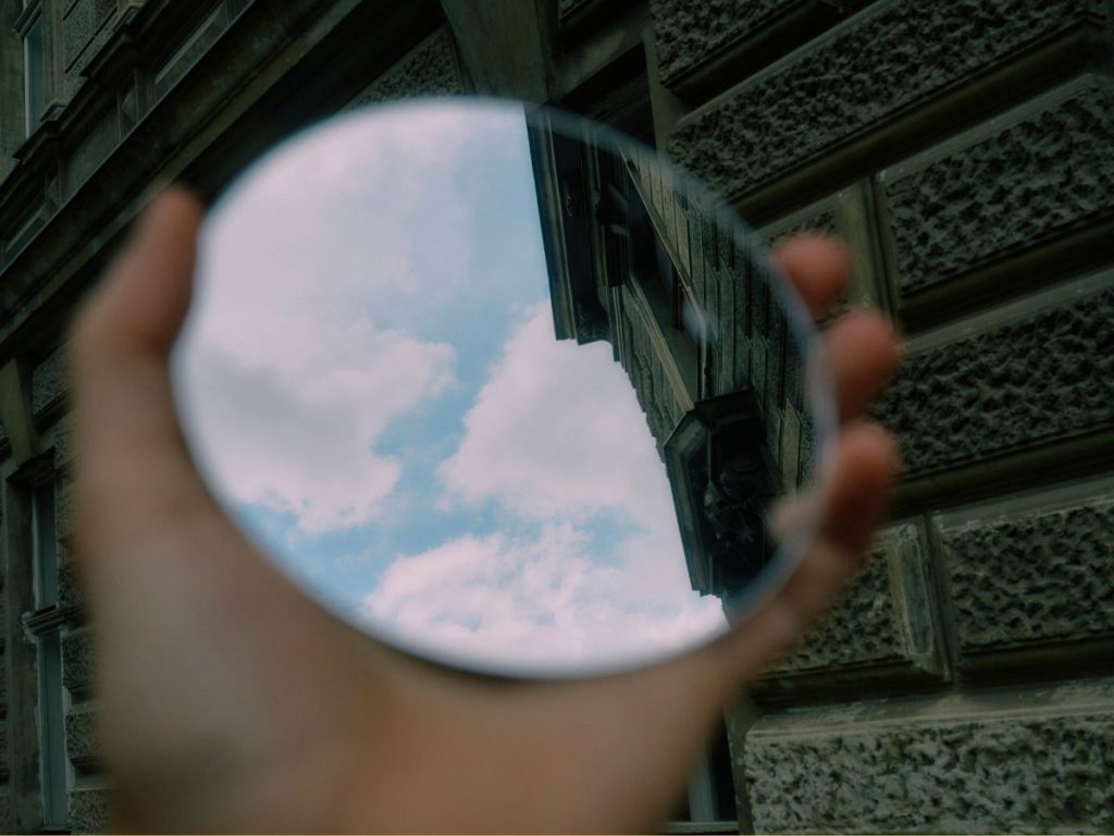 Das Foto zeigt eine Hand, die einen runden Spiegel nach oben hält. In dem Spiegel, sieht man die ein Stück des blauen Himmels.