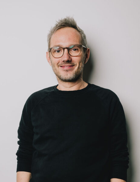 Ein weisser Mann mit kurzen Haaren und Brille blickt in die Kamera. Er trägt einen schwarzen Pullover.