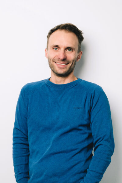 Ein weisser Mann mit kurzen Haaren blickt in die Kamera. Er trägt einen blauen Pullover.