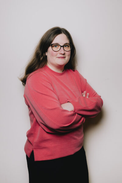 Constanze hat lange brauen Haare und trägt eine Brille. Ihr Oberteil ist hellrot, die Arme hält sie verschränkt