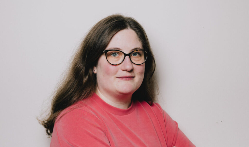 Constanze hat lange brauen Haare und trägt eine Brille. Ihr Oberteil ist hellrot, die Arme hält sie verschränkt