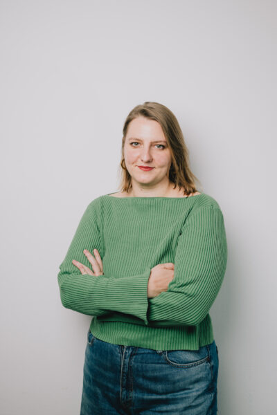 Hannah trägt ein grünes Oberteil und hat blonde Haare. Ihr Hände sind vor der Brust verschränkt.