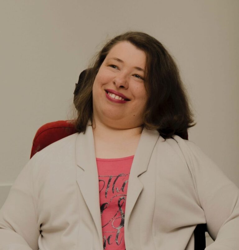 Leonie Schüler schaut in lachend in die Kamera. Sie trägt einen beigen Blazer und pinken Lippenstift. Sie nutzt einen Rollstuhl.