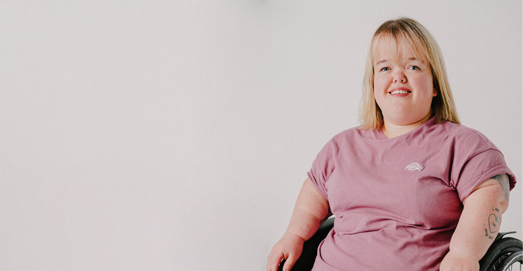 Franziska Lammers schaut mit freundlichem Blick in die Kamera. Sie trägt ein rosarotes T-Shirt und hat blonde Haare. Sie benützt einen Rollstuhl.