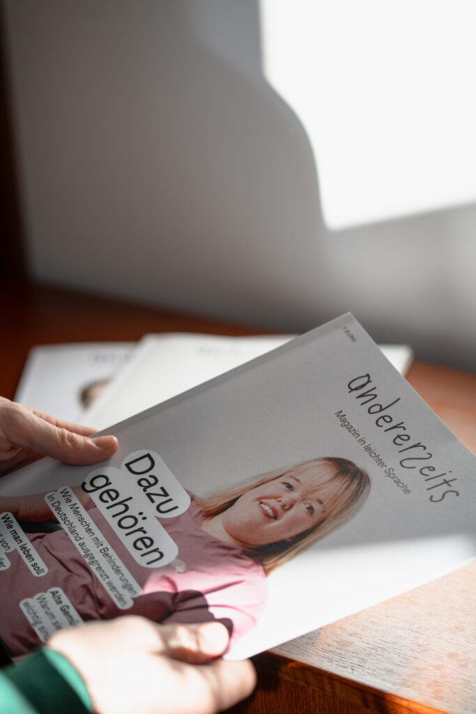 Eine Person hält das Magazin andererseits in der Hand. Auf dem Cover ist eine junge Frau in einem rosa Oberteil zu sehen. Der Titel der Ausgabe lautet "Dazugehören".