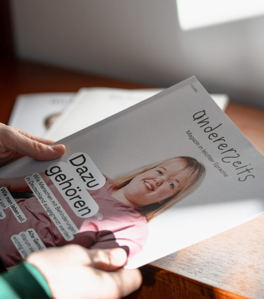 Eine Person hält das Magazin andererseits in der Hand. Auf dem Cover ist eine junge Frau in einem rosa Oberteil zu sehen. Der Titel der Ausgabe lautet "Dazugehören".