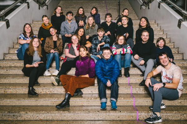 Menschen mit und ohne Behinderungen sitzen auf einer Treppe und schauen in die Kamera.