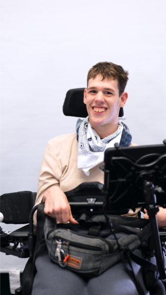 Man sieht ein junger Mann mit einem weißen Halstuch. Er sitzt in einem Elektro- Rollstuhl und lächelt in die Kamera