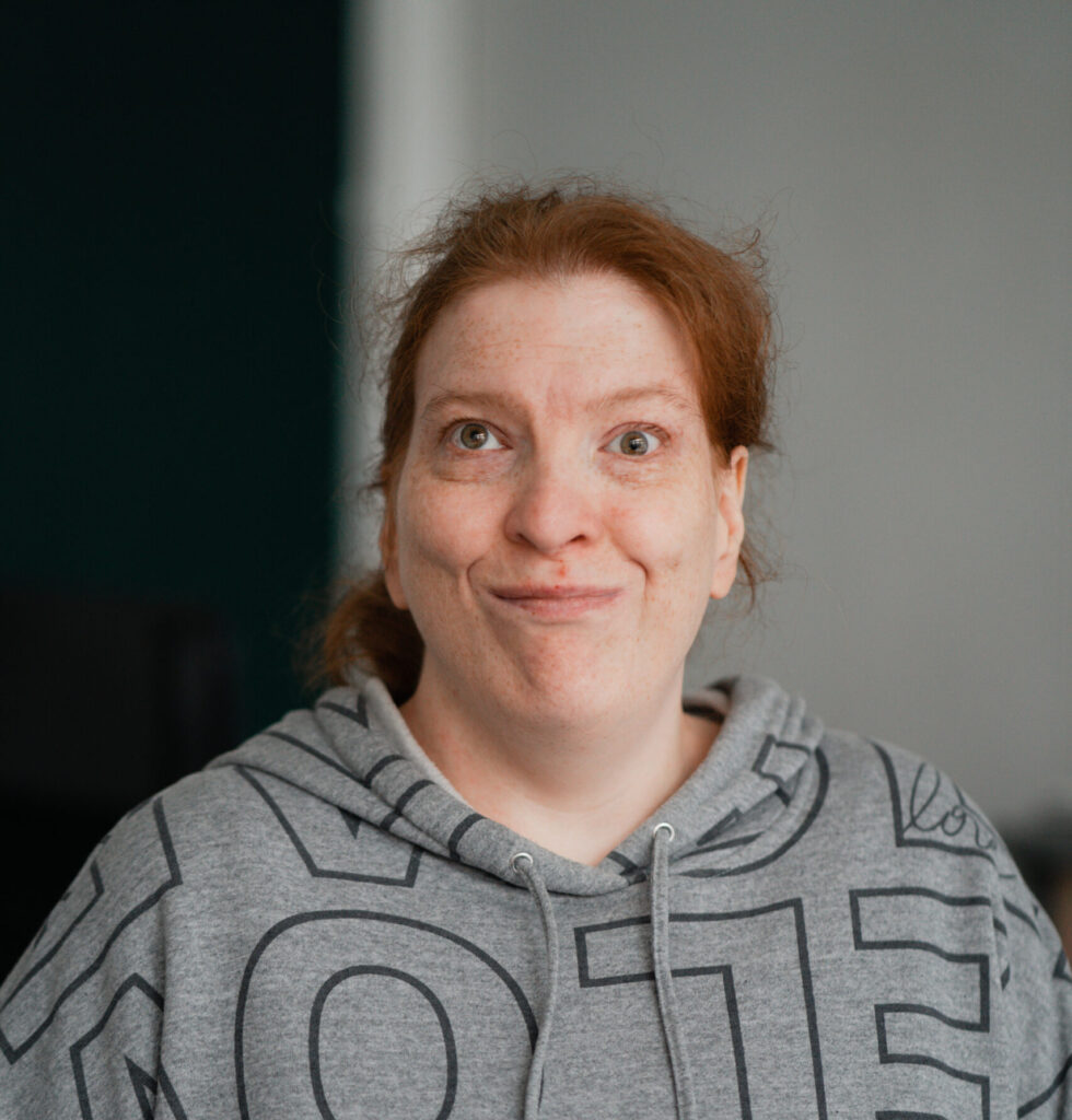 Lena schaut mit freundlichen Blick in die Kamera. Sie trägt einen grauen Pullover und hat rot-blonde Haare.