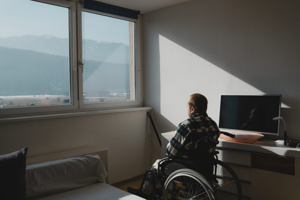 Anton sitzt in mit dem Rücken zur Kamera in einem Rollstuhl. Er schaut aus einem Fenster. Draußen sieht man Berge und einen blauen Himmel.