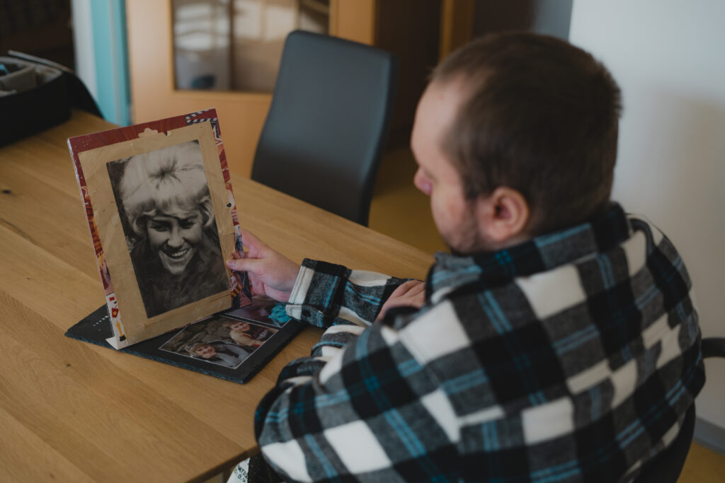 Anton sitzt an einem Tisch und hält ein Foto in der Hand. Darauf sieht. man die schwarz-weiß Aufnahme einer Frau. Er schaut es sich aufmerksam an. Auf dem Tisch liegen weitere Bilder.