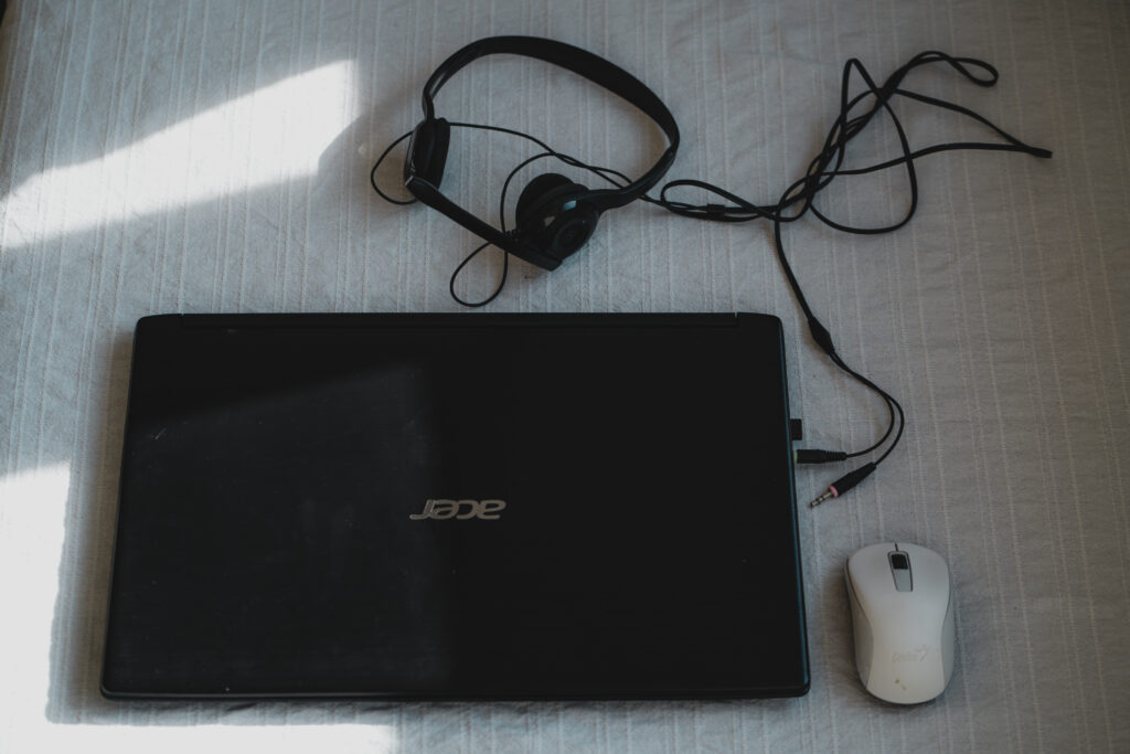 Auf dem Bild liegt ein schwarzer Laptop auf einem hellen Stoff. Daneben befindet sich eine weiße Computermaus und ein schwarzes Headset mit Kabel. Sonnenlicht fällt auf einen Teil des Stoffes und wirft Schatten.