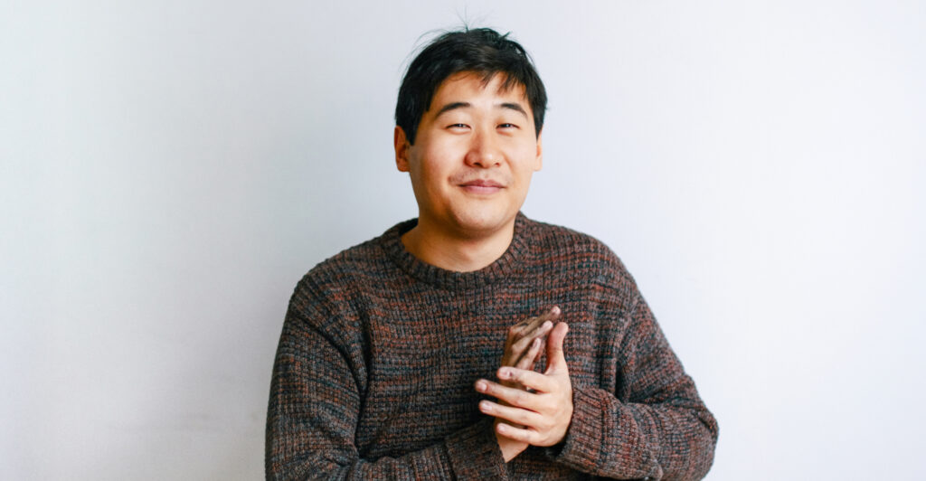 Auf diesem Foto sieht man den Künstler Juewen Zhang. Er sieht mit freundlichem Blick in die Kamera. Er trägt einen braunen Pullover.