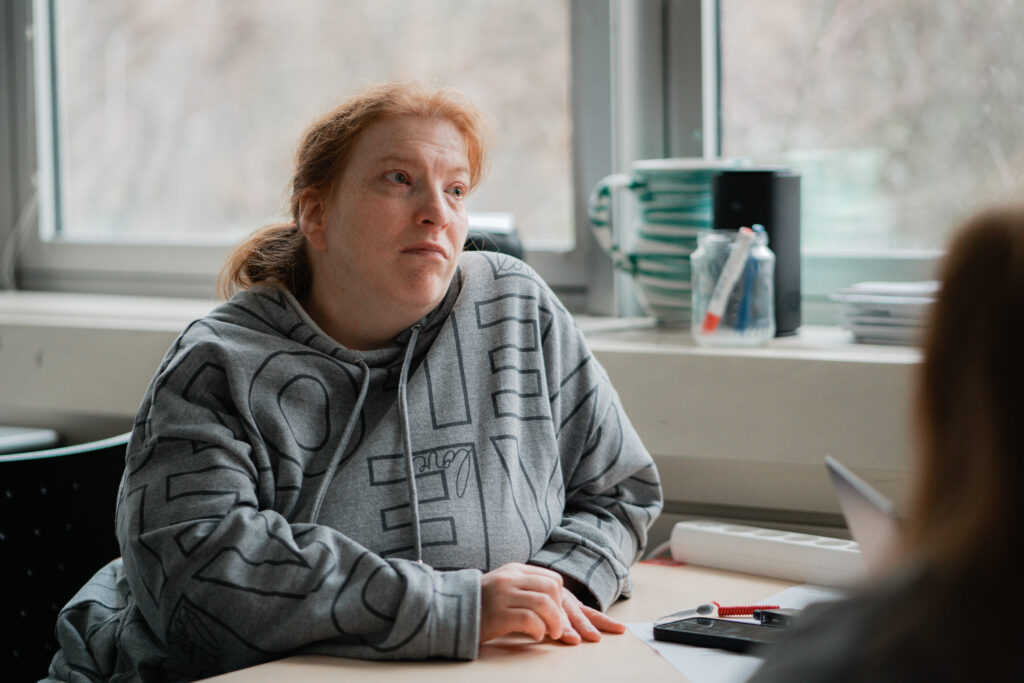 Lena sitzt an einem Tisch. Sie trägt einen grauen Hoodie mit Mustern. Sie schaut nachdenklich und hört jemandem zu. Im Hintergrund sieht man ein Fenster, eine Tasse und einige Unterlagen.