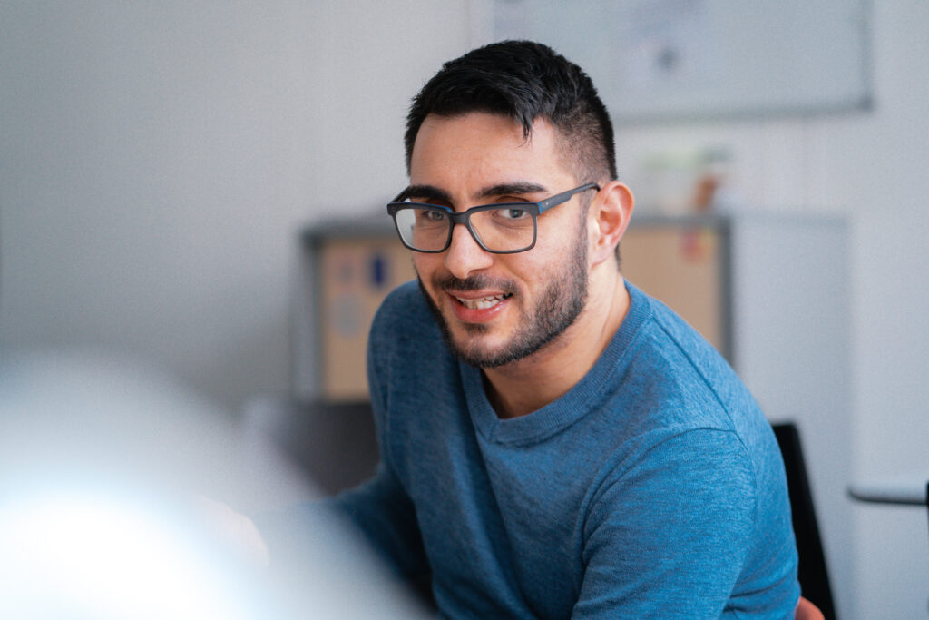 Volkan sitzt an einem Tisch. Er schaut mit ernsten Blick an der Kamera vorbei. Er trägt eine Brille und hat kurze schwarze Haare.