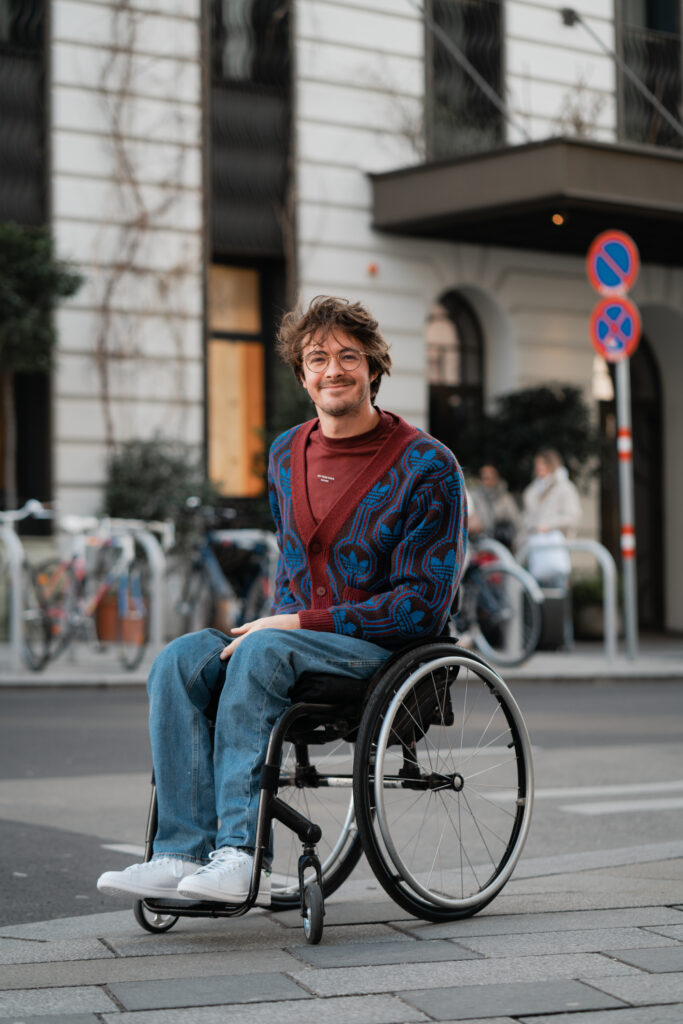 Nico sitzt in einem Rollstuhl und schaut mit freundlichen Blick in die Kamera. Er braune Haare und trägt eine Brille. Er ist draußen in der Stadt. Hinter ihm sieht man Fahrräder und ein Gebäude.