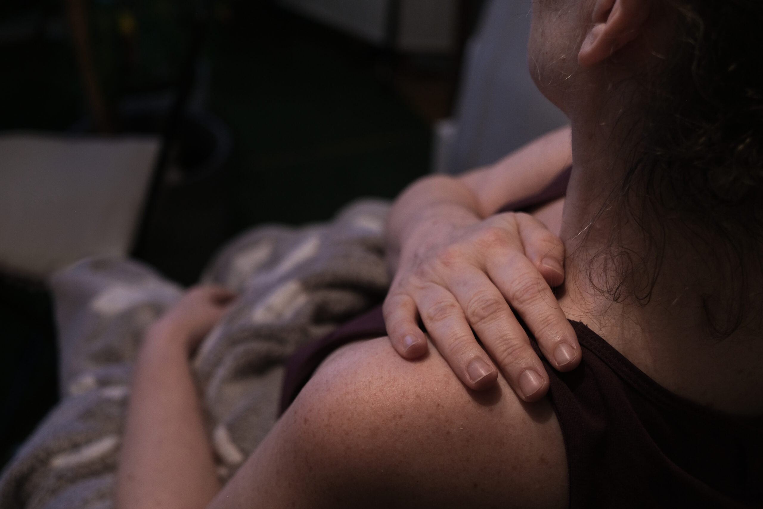 Claudia Turetschek sitzt auf einem Stuhl und hält sich mit einer Hand die Schulter. Sie trägt ein dunkles Oberteil. Eine Decke liegt über ihren Beinen. Die Umgebung ist gedimmt beleuchtet.