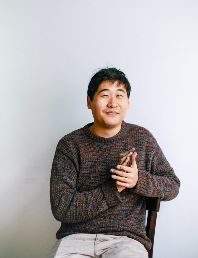 Auf diesem Foto sieht man den Künstler Juewen Zhang. Er sieht mit freundlichem Blick in die Kamera. Er trägt einen braunen Pullover.