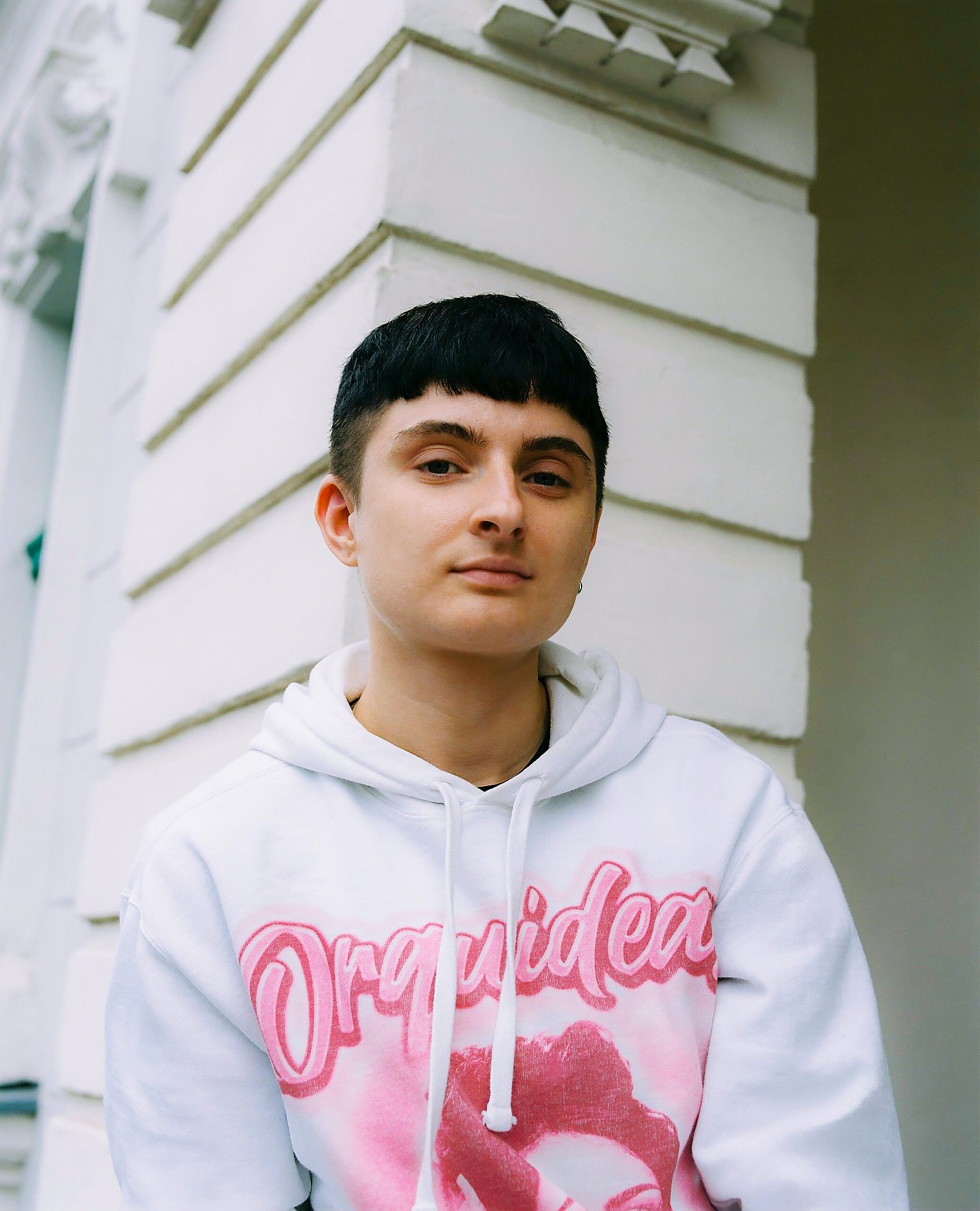 Eine Person mit kurzen schwarzen Haaren schaut mit ernstem Blick in die Kamera. Sie trägt einen weißen Pullover. Die Person steht vor einem weißen Gebäude.
