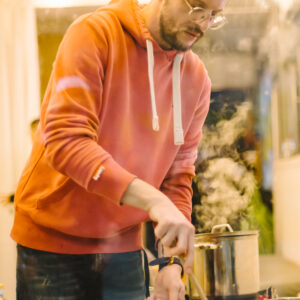 Ein Mann steht vor einer Herdplatte. Er kocht und rührt in einer Pfanne. Der Mann trägt eine Brille und einen roten Pullover.