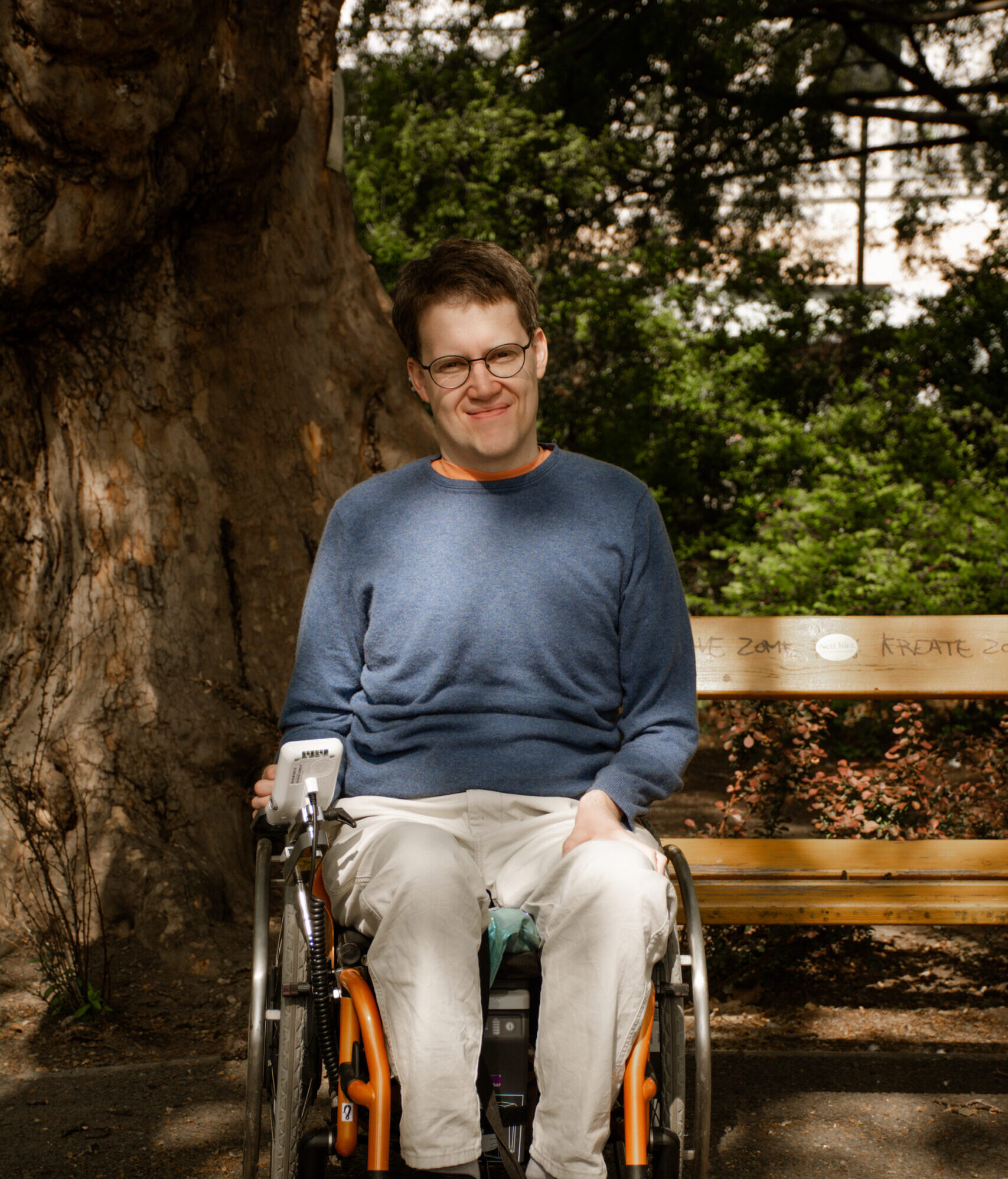 Tobias Schörghofer schaut in die Kamera. Er benützt einen Rollstuhl und trägt einen blauen Pullover und eine beige Hose. Im Hintergrund steht eine Parkbank und ein Baum.