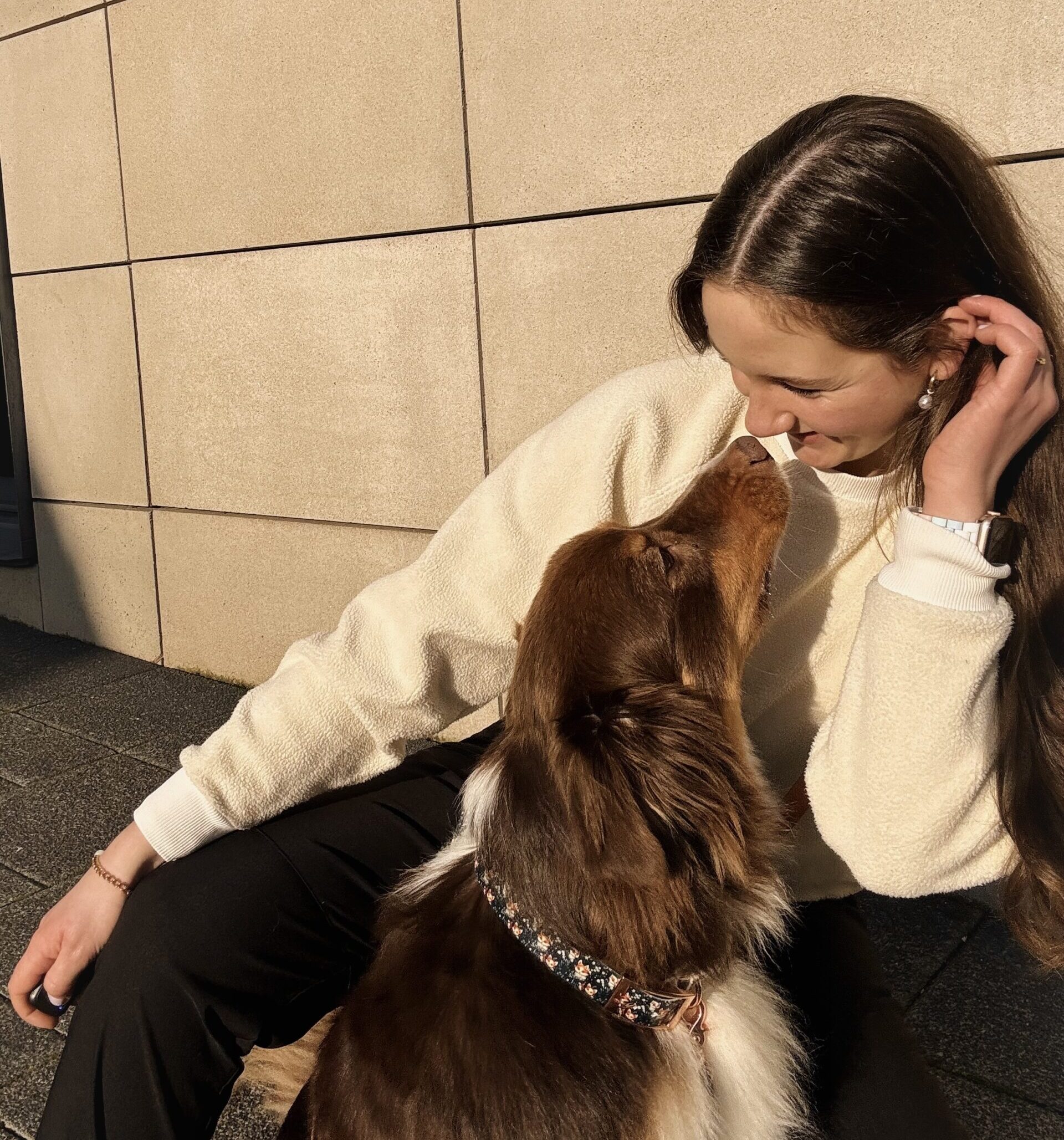 Auf dem Foto sieht man ein Mädchen, das auf dem Boden sitzt und nach unten lächelt. Ihr Hund Bailey sitzt unter ihr und schaut nach oben in ihr Gesicht.