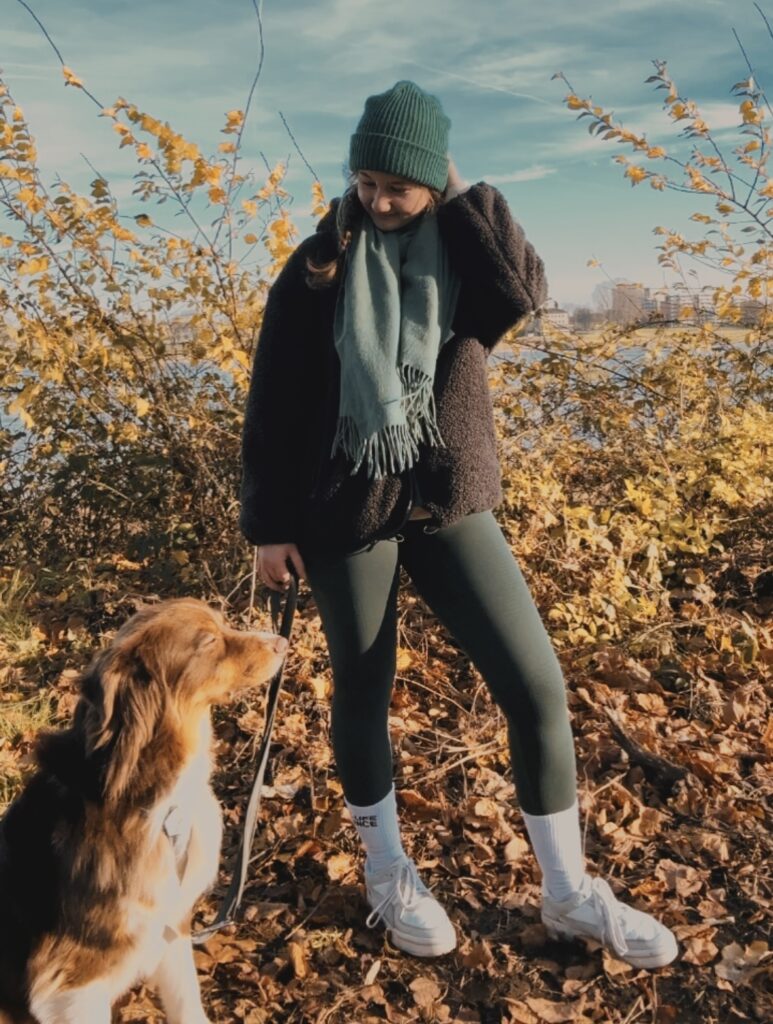 Ein Mädchen trägt warme Kleidung, eine Jacke, eine Mütze und einen Schal. In der rechten Hand hält sie eine Hundeleine. In der linken Ecke des Bildes sitzt ihr Hund Bailey. Die sind in der Natur.