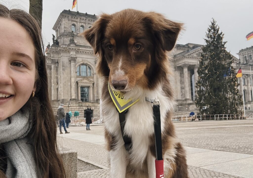 Auf dem Foto sieht man das halbe Gesicht von einem jungen Mädchen auf der linken Seite. Sie lacht in die Kamera und hat lange braune Haare. Rechts daneben steht ihr Hund Bailey und schaut auch in die Kamera. Der Hund trägt ein Halsband auf dem "Ausbildung" steht.