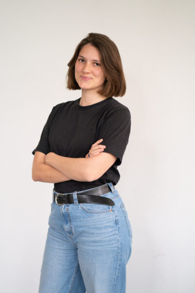Kristina trägt ein schwarzes Obteril und eine helle Jeans. Sie hat schulterlange Haare und verschränkte arme.