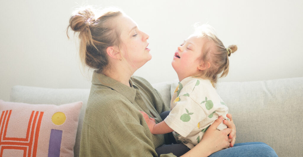 Das Bild zeigt Valerie und Emil auf einer Couch. Valerie hält Emil im Arm, beide schauen sich mit dem Gesicht zugewandt an. Die Farben sind hell und warm.
