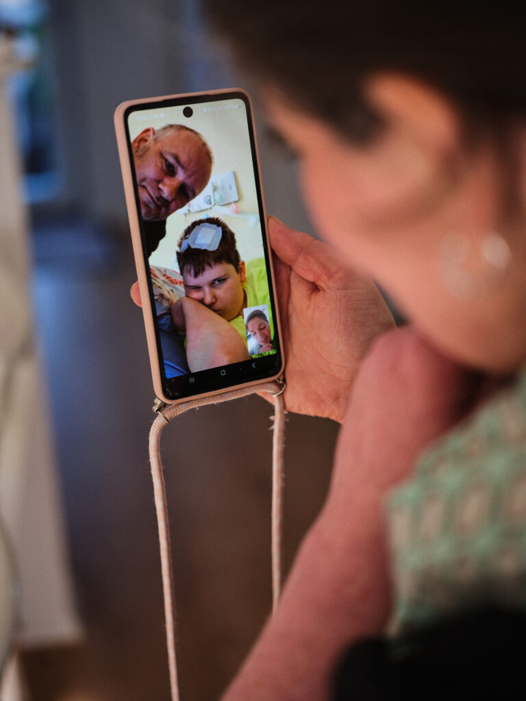 Das Foto zeigt eine Person die ein Handy in der Hand hält. Sie telefoniert via Video mit ihrer Familie. Auf dem Bildschirm sieht man einen Mann und einen Jungen mit einem Pflaster auf dem Kopf.