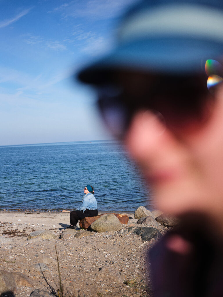 Das Foto zeigt eine unscharfe Person auf der rechten Seite des Bildes. Im Hintergrund sieht man das Meer. Am Strand sitzt eine Person und genießt die Sonne.