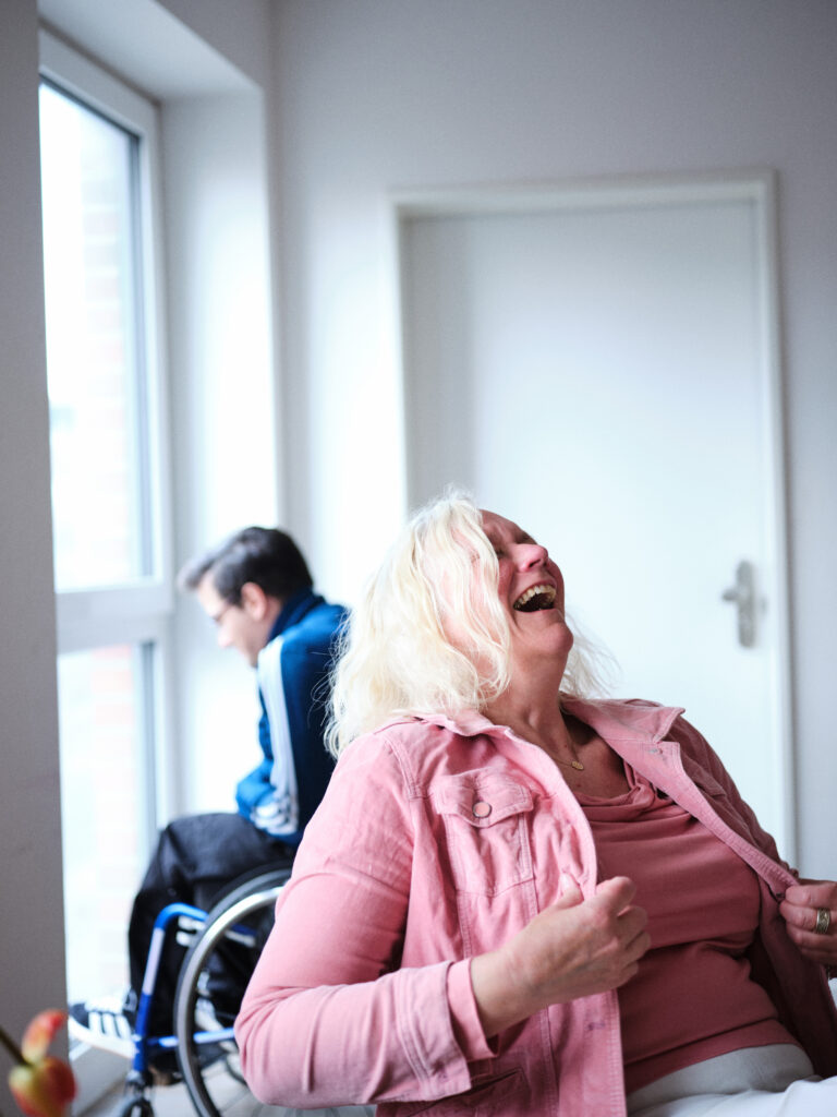 Astrid sitzt auf einem Stuhl und lacht freudig. Sie trägt eine pinke Bluse. Dahinter ist ihr Sohn Anton. Er benützt einen Rollstuhl und schaut aus einem Fenster nach draußen.