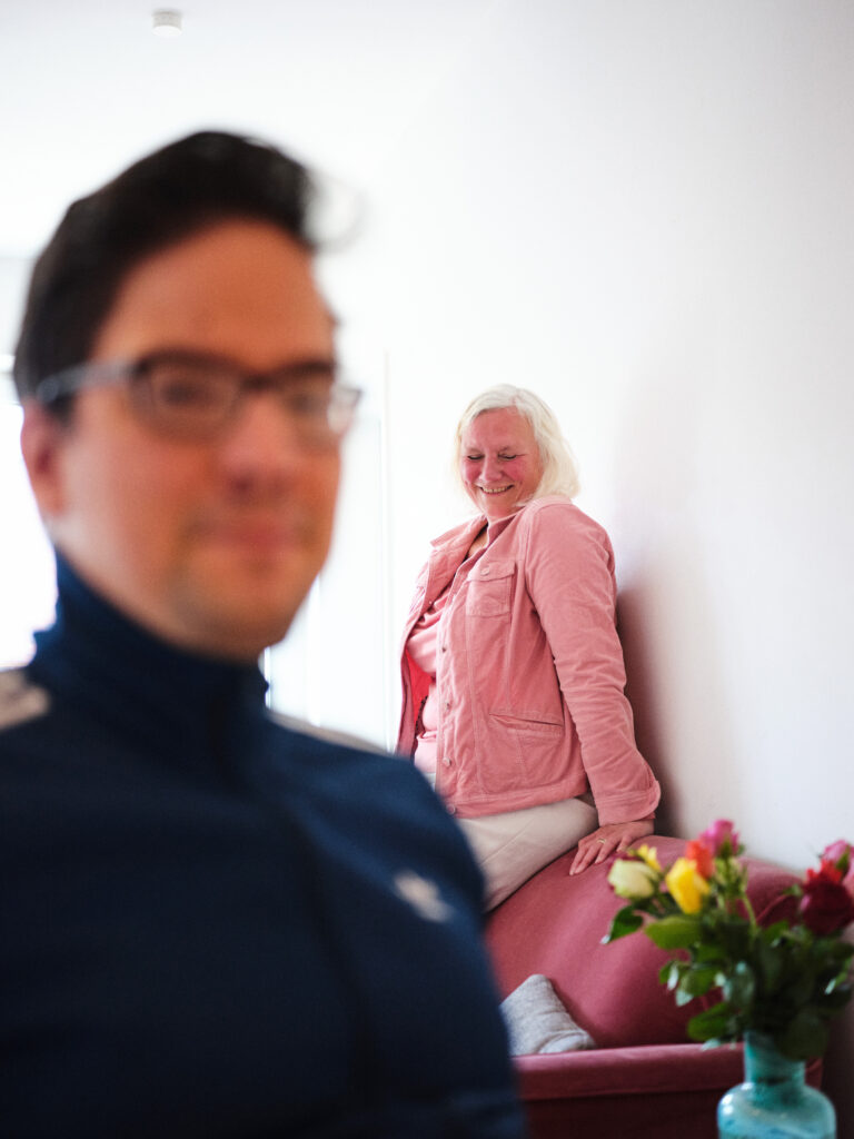 Auf dem Bild sieht man links Antons Gesicht unscharf. Er hat schwarze kurze Haare und trägt eine Brille. Dahinter auf der rechten Seite sitzt Astrid auf einem Sofa. Sie lacht mit geschlossenen Augen. Davor steht eine Vase mit Rosen.