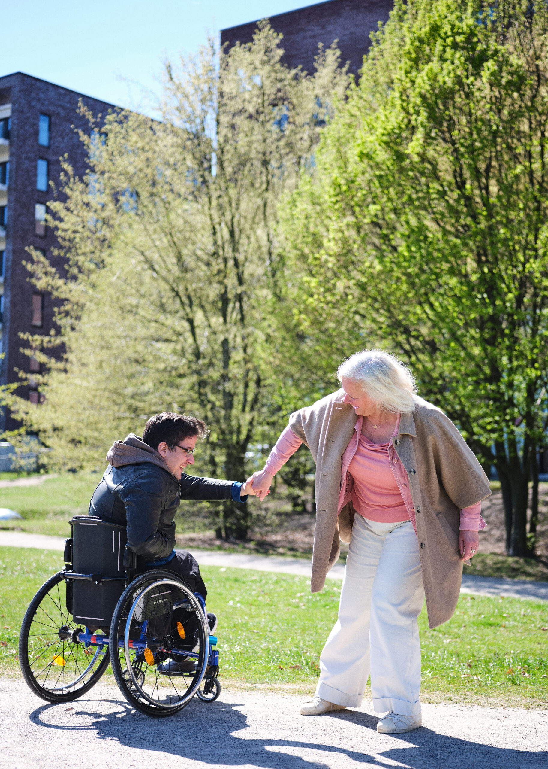 Das Foto zeigt Astrid mit ihrem Sohn Anton. Sie sind in einem Park. Er nutzt einen Rollstuhl. Sie halten sich an den Händen und lachen aneinander an.
