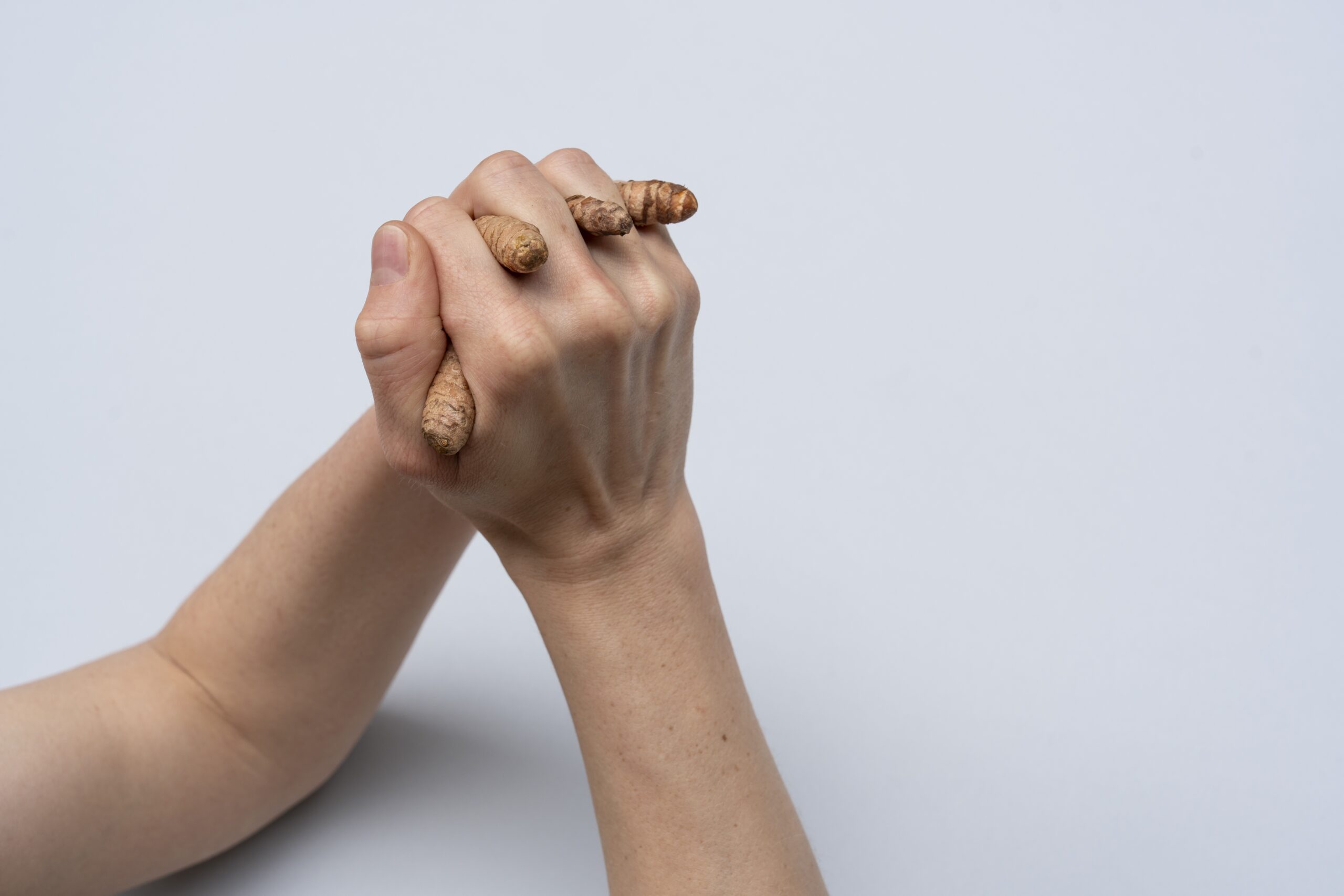 Zwei Hände sind einander verhakt. Die Finger der linken Hand sind Kurkuma-Knollen. Der Hintergrund ist weiß.
