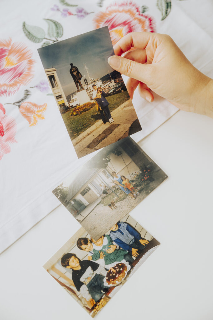 Das Bild zeigt analoge Fotos auf einem Tisch. Rechts oben hält eine Hand ein Foto hoch. Auf den Bildern sieht man mehrere Menschen.