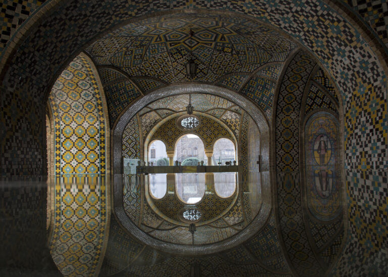 Auf dem Bild ist ein Raum. Er hat viele Rundbögen und eine spiegelnde Fläche auf dem Boden. Es ist ein Teil des Saadabad Palasts in Teheran. Die Decke ist mit Mustern verziert. In der Mitte sieht man Bögen mit Blick nach draußen.