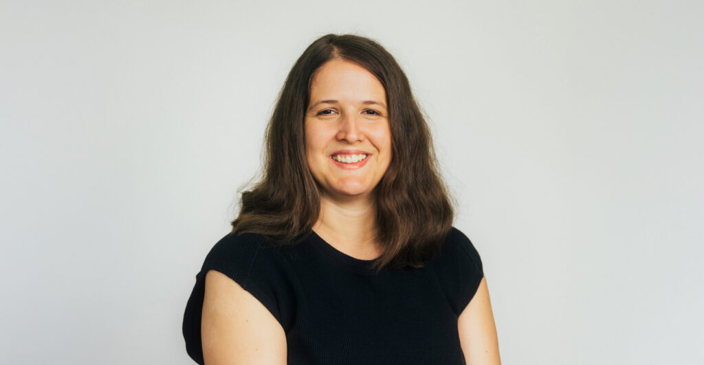 Das Foto zeigt eine junge Frau. Sie lächelt in die Kamera. Sie hat braune lange Haare und trägt ein schwarzes T-Shirt.