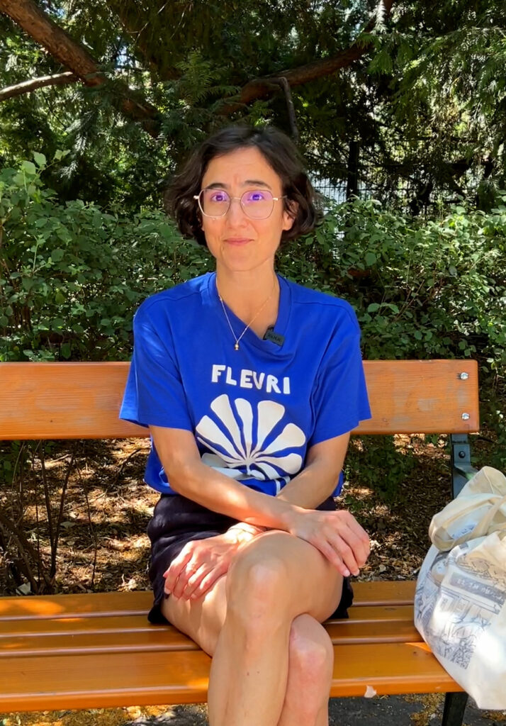 Eine Frau schaut mit ernstem Blick in die Kamera. Sie trägt eine Brille und hat kurze braune Haare. Sie sie trägt ein blaues T-Shirt und sitzt auf einer Parkbank.
