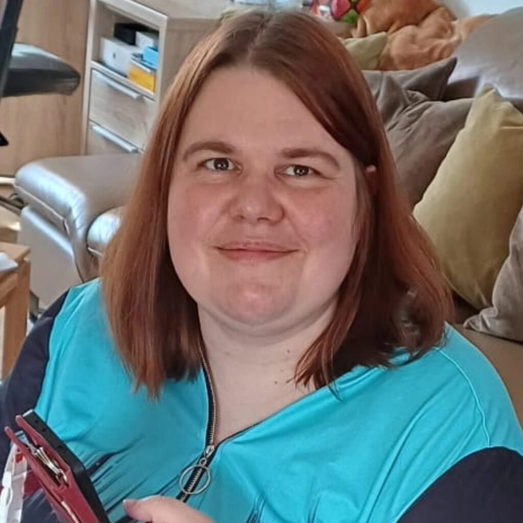 Eine junge Frau schaut mit freundlichen Blick in die Kamera. Sie hat braune schulterlange Haare. Sie trägt eine blaue Jacke. Im Hintergrund sieht man ein Sofa.