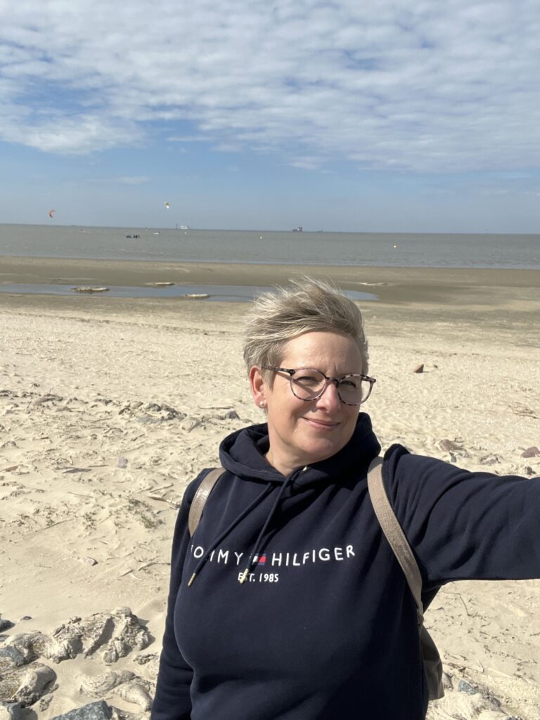 Eine Frau schaut mit zusammengekniffenen Augen in die Kamera. Sie hat kurze blonde Haare und trägt eine Brille. Sie steht an einem Strand, im Hintergrund sieht man das Meer.
