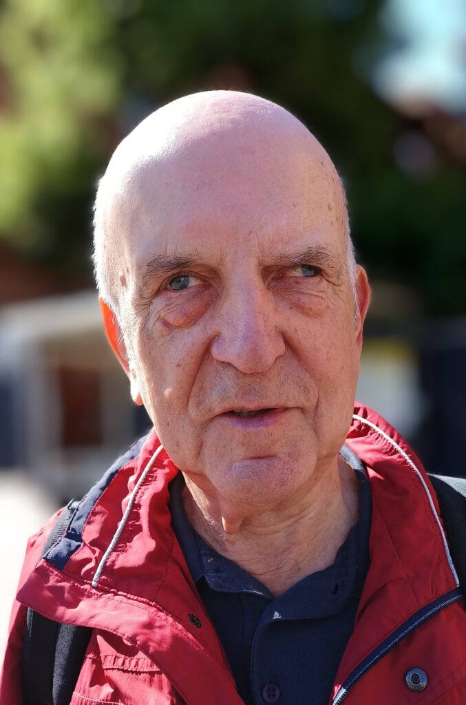 Ein älterer Mann schaut mit ernstem Blick geradeaus. Er hat eine Glatze und trägt eine rote Jacke.