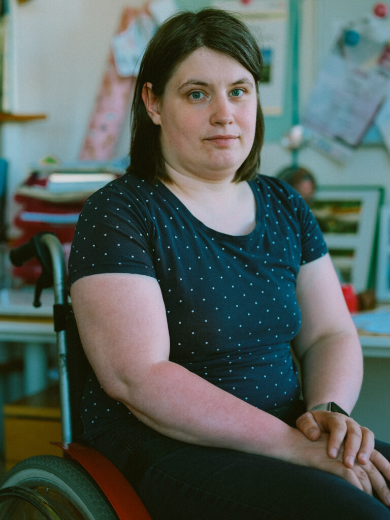 Eine junge Frau sieht mit ernstem Blick in die Kamera. Sie hat kurze braune Haare und trägt ein T-Shirt. Sie benutzt einen Rollstuhl.