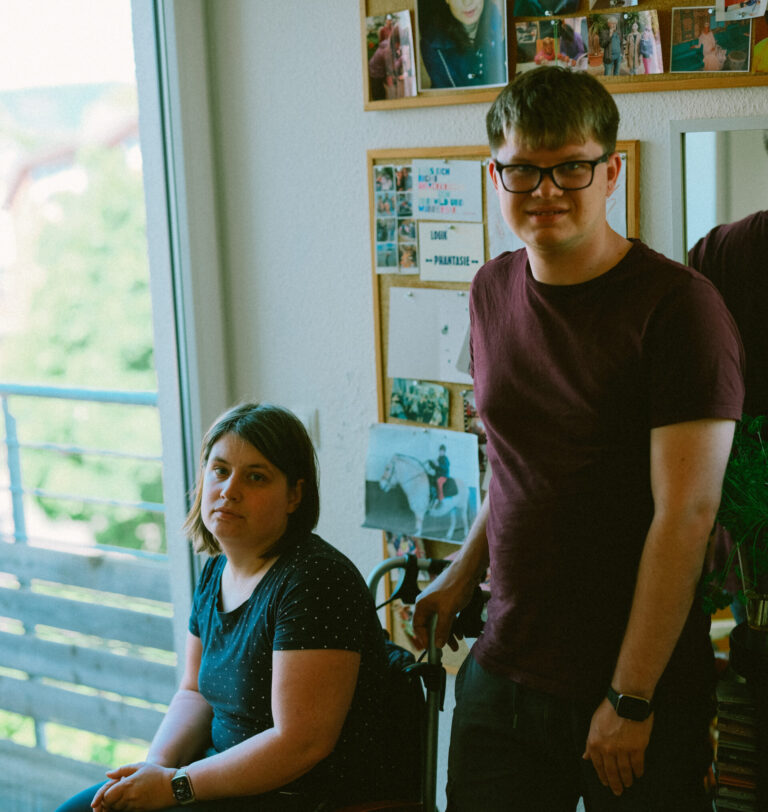 Zwei Personen schauen mit ernsten Blick in die Kamera. Die linke Person ist eine junge Frau, sie benutzt einen Rollstuhl und trägt ein T-Shirt. Rechts daneben steht ein junger Mann. Er trägt eine Brille und hat kurze Haare.